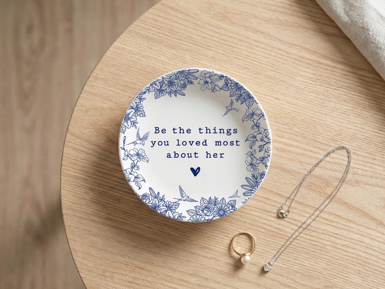 Może przedstawiać: Okrągła biała ceramiczna miseczka z niebieską kwiatową obw&oacute;dką z małymi ptakami oraz napisem "Be the things you loved most about her" wydrukowanym na środku nad małą niebieską ikoną serca, w towarzystwie złotego pierścionka i srebrnego naszyjnika.