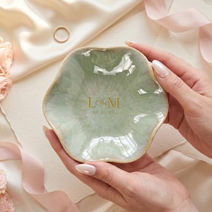 May include: A sage green ceramic ring dish with a scalloped, flower-like edge. The center features gold lettering with the initials L and M separated by a small heart symbol, followed by the date 08.21.25 printed in a delicate gold font.