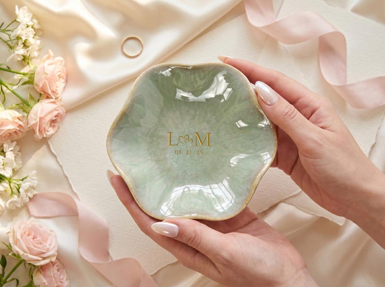 May include: A sage green ceramic ring dish with a scalloped, flower-like edge. The center features gold lettering with the initials L and M separated by a small heart symbol, followed by the date 08.21.25 printed in a delicate gold font.
