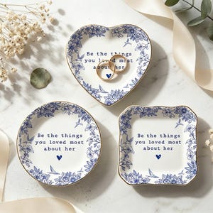 Może przedstawiać: Trzy białe ceramiczne miseczki na drobiazgi ze złotymi obrzeżami i niebieskimi wzorami kwiatowymi. Każda miseczka zawiera napis "Be the things you loved most about her" oraz małe niebieskie serce. Dwa złote pierścionki leżą w miseczce w kształcie serca.