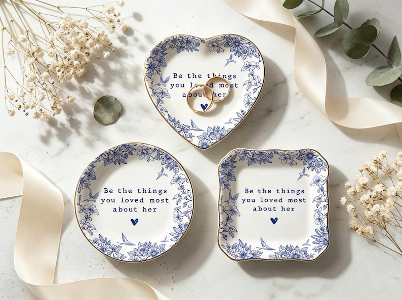 Może przedstawiać: Trzy białe ceramiczne miseczki na drobiazgi ze złotymi obrzeżami i niebieskimi wzorami kwiatowymi. Każda miseczka zawiera napis "Be the things you loved most about her" oraz małe niebieskie serce. Dwa złote pierścionki leżą w miseczce w kształcie serca.