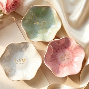 May include: Three flower-shaped ceramic trinket dishes in white, pale green, and pink with a glossy finish. Each dish features gold-colored custom lettering and dates. The white dish displays L&M 08.21.25, the green dish shows k&f 07.09.2025, and the pink dish shows h&b 08.22.2026.
