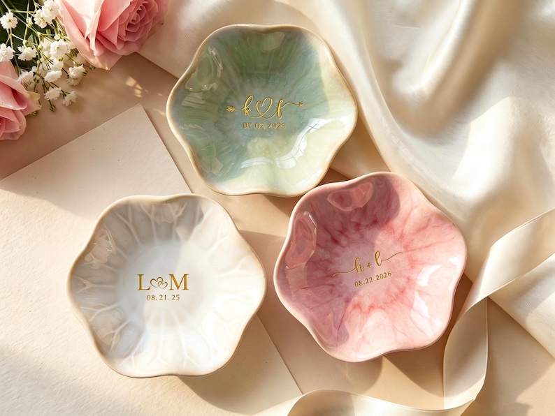 May include: Three flower-shaped ceramic trinket dishes in white, pale green, and pink with a glossy finish. Each dish features gold-colored custom lettering and dates. The white dish displays L&M 08.21.25, the green dish shows k&f 07.09.2025, and the pink dish shows h&b 08.22.2026.