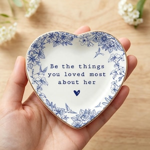 Może przedstawiać: Biała ceramiczna miseczka w kształcie serca z niebieskim kwiatowym wzorem na obrzeżach oraz nadrukowanym na środku tekstem "Be the things you loved most about her", kt&oacute;remu towarzyszy mała niebieska ikona serca pod tekstem.