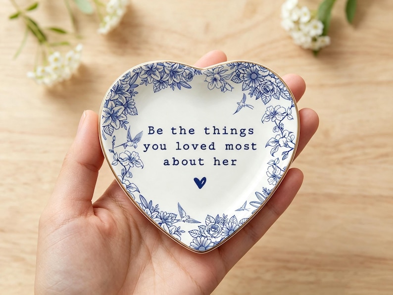 Może przedstawiać: Biała ceramiczna miseczka w kształcie serca z niebieskim kwiatowym wzorem na obrzeżach oraz nadrukowanym na środku tekstem "Be the things you loved most about her", kt&oacute;remu towarzyszy mała niebieska ikona serca pod tekstem.