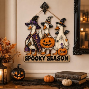 May include: A decorative wall hanging featuring three white geese dressed for Halloween. One wears a purple star-patterned witch hat and cape, one holds a jack-o'-lantern, and one wears a ghost sheet. The text SPOOKY SEASON is printed below.