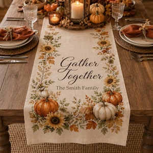 May include: A beige fabric table runner featuring a printed autumn design with sunflowers, orange and white pumpkins, and fall foliage. The text "Gather Together" and "The Smith Family" is centered in black script and serif lettering on the runner.
