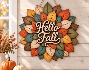May include: A circular wall hanging featuring layered wooden leaves in shades of orange, yellow, brown, and teal. The center displays the text Hello Fall in a light-colored script font. The piece is suspended by a rope loop at the top.