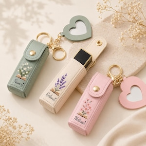 May include: A collection of personalized lipstick holders with keychains. The holders are in pastel colors: sage green, cream, and pink. Each case features floral embroidery and a name. A heart-shaped mirror charm is attached to two of the keychains.