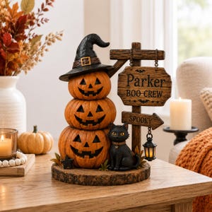 Puede incluir: Una figura decorativa de mesa con tres calabazas naranjas apiladas, una de ellas con un sombrero de bruja negro. Al lado hay un gato negro y un poste de madera con el texto Parker BOO CREW y un letrero más pequeño que dice SPOOKY.