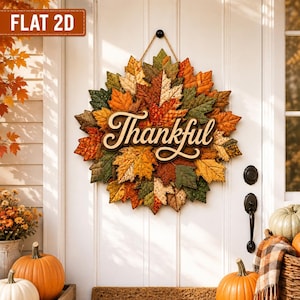 May include: A circular wall hanging featuring a dense arrangement of fabric autumn leaves in shades of orange, yellow, brown, and green. A wooden cutout script sign with the word Thankful is centred over the leaves, suspended by a thin cord.