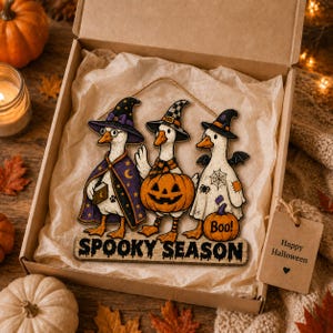 May include: A decorative wooden sign featuring three white geese dressed in Halloween costumes, including witch hats and capes, with a jack-o'-lantern and the text "SPOOKY SEASON" at the bottom, packaged in a cardboard gift box with a tag reading "Happy Halloween".