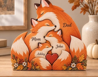 May include: A decorative wooden fox family figurine featuring four foxes huddled together with closed eyes. The foxes are labeled with the names Dad, Mom, John, and Ashley. A small red heart is positioned between the two smaller foxes, and the base is decorated with painted autumn leaves, berries, and white flowers.