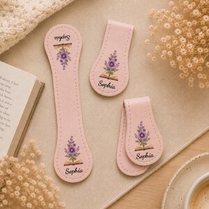 May include: Three pink leather book accessories with a floral design and the name "Sophia". The accessories include a long bookmark, and two magnetic page holders. The floral design features purple flowers and an open book.