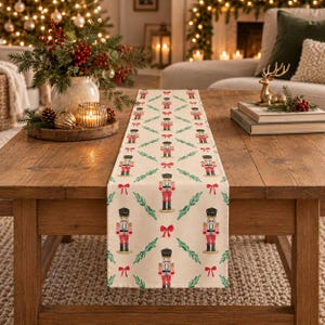 May include: A cream-colored table runner featuring a repeating pattern of nutcracker figures in red and black uniforms, interspersed with green sprigs and small red bows, laid flat across a wooden table surface.