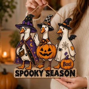May include: A wooden hanging sign featuring three geese dressed in Halloween costumes. One wears a purple star-patterned cloak and hat, one holds a jack-o'-lantern, and one wears a ghost sheet with a spider web. The sign reads SPOOKY SEASON.