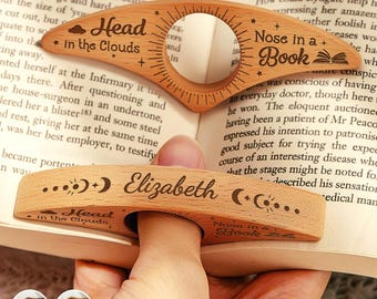 May include: Wooden thumb page holders with engraved text. One has "Head in the Clouds" and "Nose in a Book" with a circular opening. The other has "Elizabeth" and celestial designs. The design can be customized.