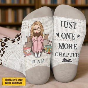May include: A pair of custom socks with a book-themed design. One sock features a cartoon illustration of a person reading with the name "Olivia". The other sock has the text "Just One More Chapter". The socks have a beige heel and toe.