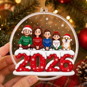 May include: A clear circular Christmas ornament featuring a cartoon illustration of two adults, two children, and a dog sitting on top of large, red, snow-capped numbers that spell out 2026. The figures wear festive sweaters and winter hats.