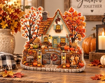 May include: A decorative tabletop house figurine featuring autumn foliage, pumpkins, and a squirrel. The structure includes a stone-style fence, a gated entrance with the text "The Martin Family," and illuminated windows set against a backdrop of orange and red trees.