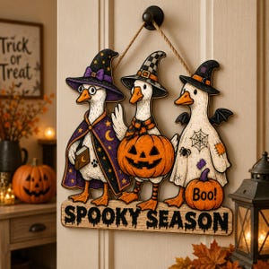May include: A hanging wooden sign featuring three white geese dressed in Halloween costumes. One wears a purple witch hat and cape, one holds a jack-o'-lantern, and one wears a ghost sheet with a spiderweb. The sign reads SPOOKY SEASON.