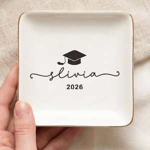 May include: A square, white ceramic trinket dish with a gold rim. The dish features a black graduation cap graphic, the name "Olivia" in cursive, and the year "2026". The dish is held by a person.