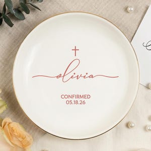 May include: A white ceramic ring dish with a gold rim, featuring a cross and the name "Olivia" in a cursive font. The dish also includes the word "CONFIRMED" and a date. The dish is surrounded by flowers and pearls.