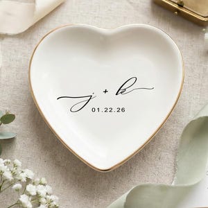 May include: A heart-shaped, white ceramic ring dish with a gold rim. The dish features the initials "J + K" and the date "01.22.26" in black script. The dish is surrounded by decorative elements, including ribbons and flowers.