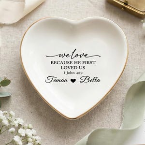 Könnte beinhalten: Eine herzförmige, weiße Keramik-Schmuckschale mit goldenem Rand. Die Schale trägt den Text "we love BECAUSE HE FIRST LOVED US 1 John 4:19 Teman ❤ Bella" in schwarzer Schrift. Dekorative Elemente sind Blumen und Bänder.
