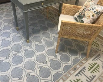 May include: A room scene featuring a gray desk with gold accents, a woven chair with a floral pillow, and two patterned rugs. The rugs have a geometric design in shades of gray, beige, and green. The desk has two drawers and is set against a wall.