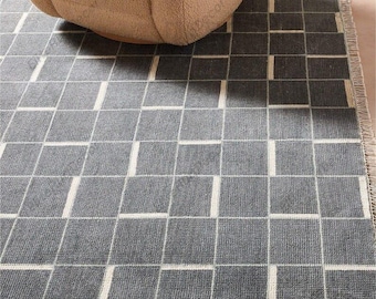 May include: A gray area rug with a geometric square pattern and white vertical lines. The rug has a fringed edge and is partially covered by a beige armchair.