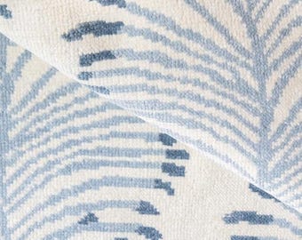May include: Close-up of a white and blue patterned rug. The rug features a repeating design of stylized blue leaves against a white background. The texture of the rug is visible, with a folded section revealing the underside.