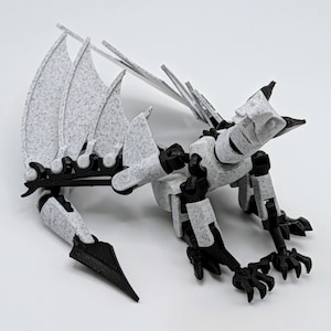 May include: A 3D-printed dragon figurine in a speckled white and black color scheme. The dragon has articulated joints, large wings, and a detailed, textured surface. The wings are a fan-like shape. The dragon is posed on a white surface.