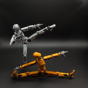 May include: Two articulated action figures in dynamic poses against a black backdrop. One figure is silver and kicking, while the other is gold and in a split. These poseable figures are designed for display or stop-motion animation.