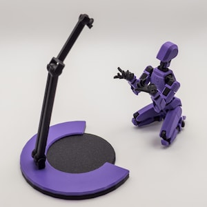 May include: A purple articulated robot figure kneels near a black and purple display stand. The robot has black joints and is posed with its hands up. The stand has a circular base and a black support arm.