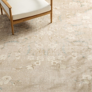 Puede incluir: Alfombra beige con estampado floral en crema, azul claro y beige. Una silla de madera con un cojín blanco está en la esquina superior izquierda. La alfombra tiene un aspecto envejecido.