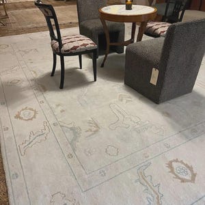 Op de afbeelding: Een lichtgekleurd vloerkleed met een gedetailleerde rand en ingewikkelde patronen. Een kleine ronde tafel en drie stoelen met een patroonbekleding omringen het vloerkleed. De stoelen zijn donker gekleurd met een mix van texturen.