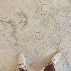 Op de afbeelding: Cr&egrave;mekleurig vloerkleed met een vervaagd, patroon in tinten lichtblauw, beige en bruin. Het vloerkleed heeft een rechthoekige vorm en een rand. Een paar witte leren instappers met goudkleurige details zijn zichtbaar.