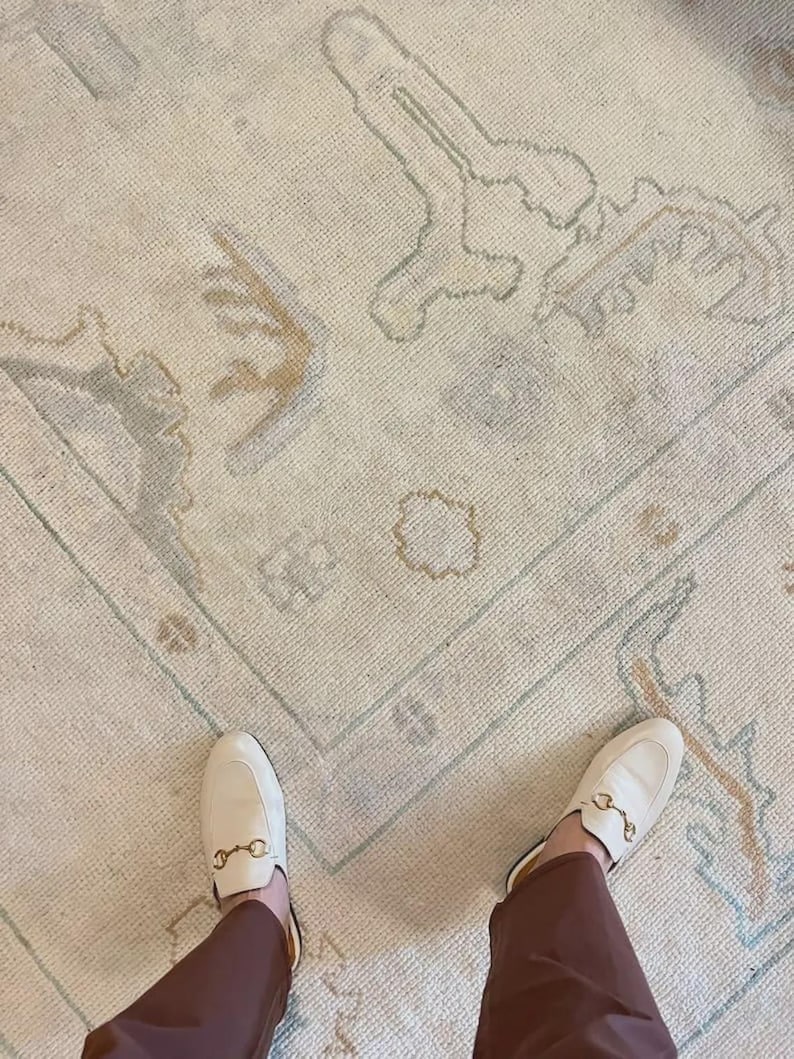 Op de afbeelding: Cr&egrave;mekleurig vloerkleed met een vervaagd, patroon in tinten lichtblauw, beige en bruin. Het vloerkleed heeft een rechthoekige vorm en een rand. Een paar witte leren instappers met goudkleurige details zijn zichtbaar.