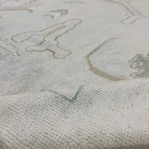 Op de afbeelding: Close-up van een cr&egrave;mekleurig tapijt met een getextureerd oppervlak. Het tapijt heeft een patroon van abstracte vormen in bruin- en lichtblauwtinten. Het tapijt is opgevouwen en onthult zijn dikke pool.