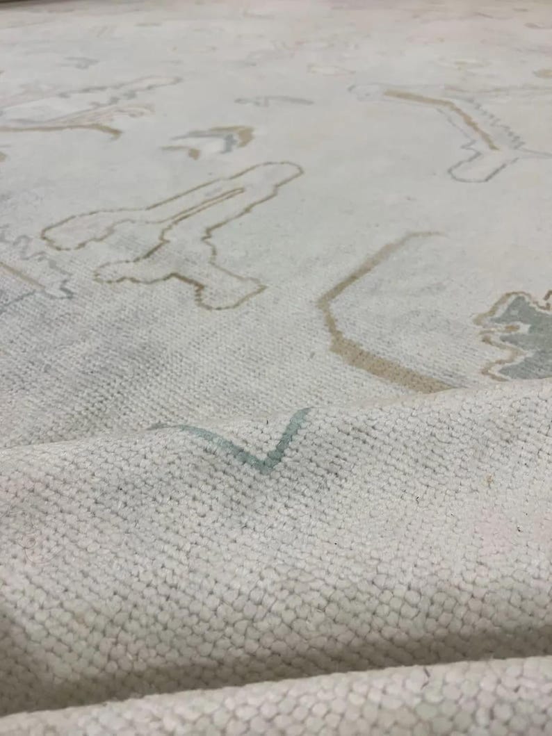 Op de afbeelding: Close-up van een cr&egrave;mekleurig tapijt met een getextureerd oppervlak. Het tapijt heeft een patroon van abstracte vormen in bruin- en lichtblauwtinten. Het tapijt is opgevouwen en onthult zijn dikke pool.