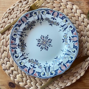 May include: A white ceramic plate with a detailed floral and geometric design in navy blue, red, and yellow. The plate is surrounded by gold-colored cutlery and rests on a woven placemat. A decorative piece for the dining table.
