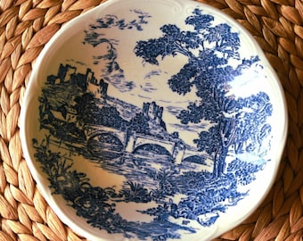 May include: A white ceramic bowl with a blue landscape design. The bowl features a detailed illustration of a bridge, castle, and trees. The rim has a scalloped edge. The bowl is set on a woven, tan surface.