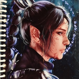 May include: Spiral-bound notebook featuring a fantasy illustration of a person with pointed ears, dark hair, and blood-like markings. The artwork is set against a dark blue background with a glowing circular design. The notebook has a white spiral binding.