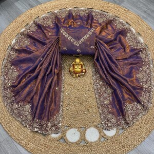 May include: A draped purple and gold sari with intricate embroidered detailing along the edges. The sari is paired with a matching blouse and a small gold Buddha statue. The sari is displayed on a round, woven jute rug.