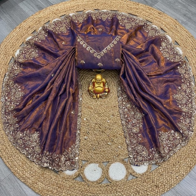 May include: A draped purple and gold sari with intricate embroidered detailing along the edges. The sari is paired with a matching blouse and a small gold Buddha statue. The sari is displayed on a round, woven jute rug.