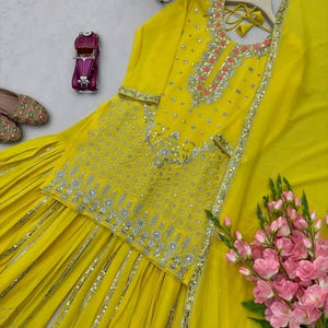 Pu&ograve; includere: Un completo ricamato giallo brillante con dettagli argentati. L'ensemble include un top decorato, pantaloni abbinati e una sciarpa fluida. L'outfit &egrave; adornato da ricami e paillettes. Sono presenti anche fiori rosa.
