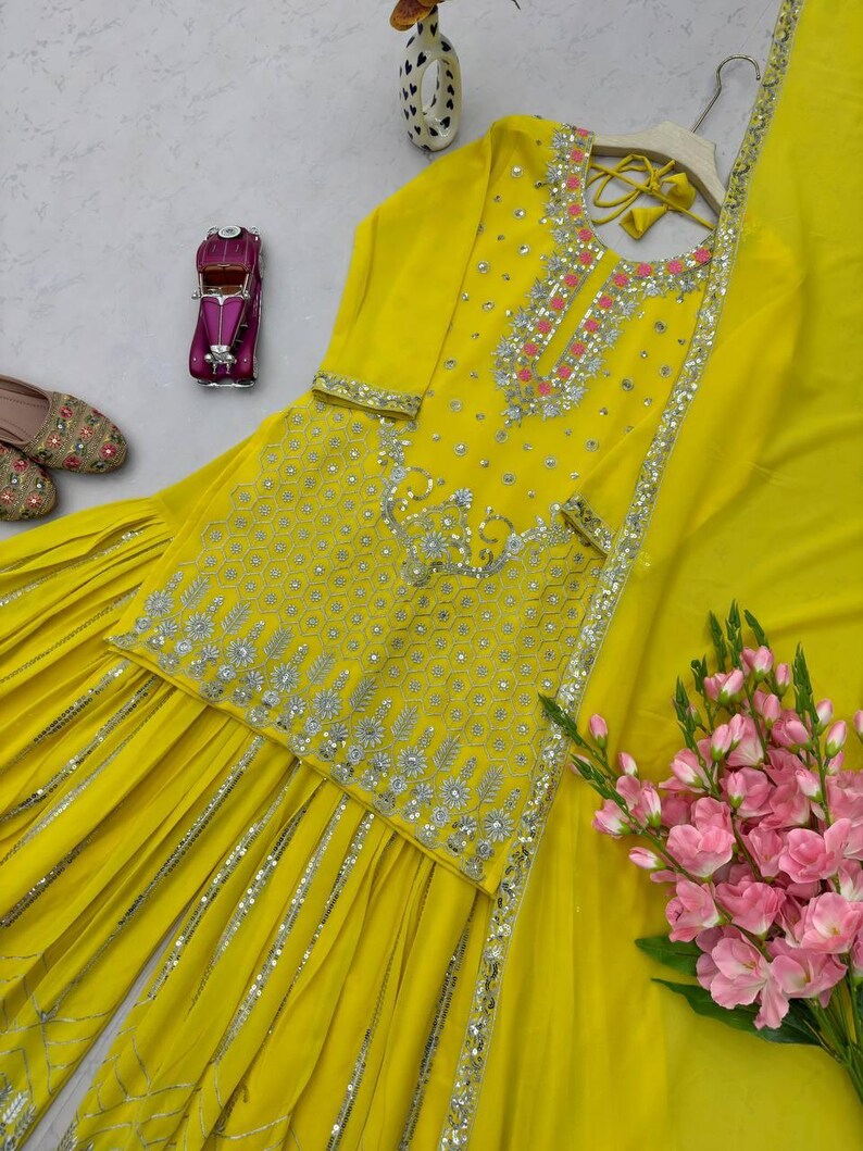 Pu&ograve; includere: Un completo ricamato giallo brillante con dettagli argentati. L'ensemble include un top decorato, pantaloni abbinati e una sciarpa fluida. L'outfit &egrave; adornato da ricami e paillettes. Sono presenti anche fiori rosa.