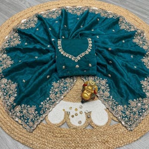 May include: Teal-colored saree with intricate gold embroidery along the borders and a matching blouse. The saree is draped over a woven mat, with a small gold Buddha statue in the foreground. The blouse features detailed embellishments around the neckline.