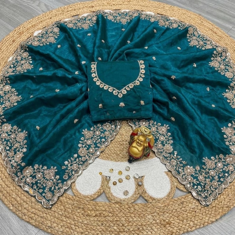 May include: Teal-colored saree with intricate gold embroidery along the borders and a matching blouse. The saree is draped over a woven mat, with a small gold Buddha statue in the foreground. The blouse features detailed embellishments around the neckline.